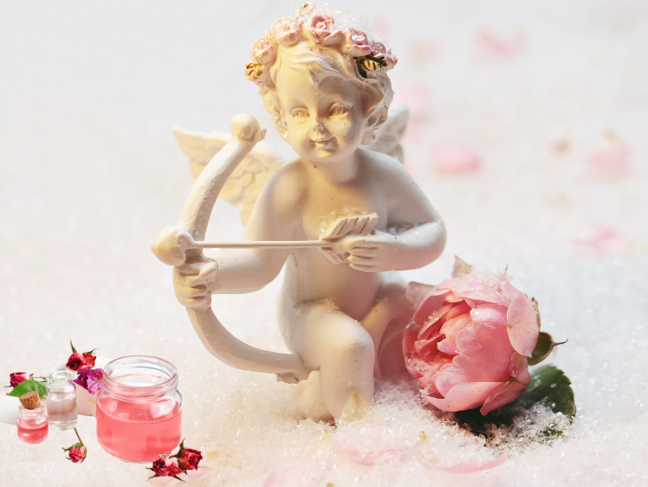 A cupid with bow and arrow, pink rose and jar of rose water, surrounded by roses